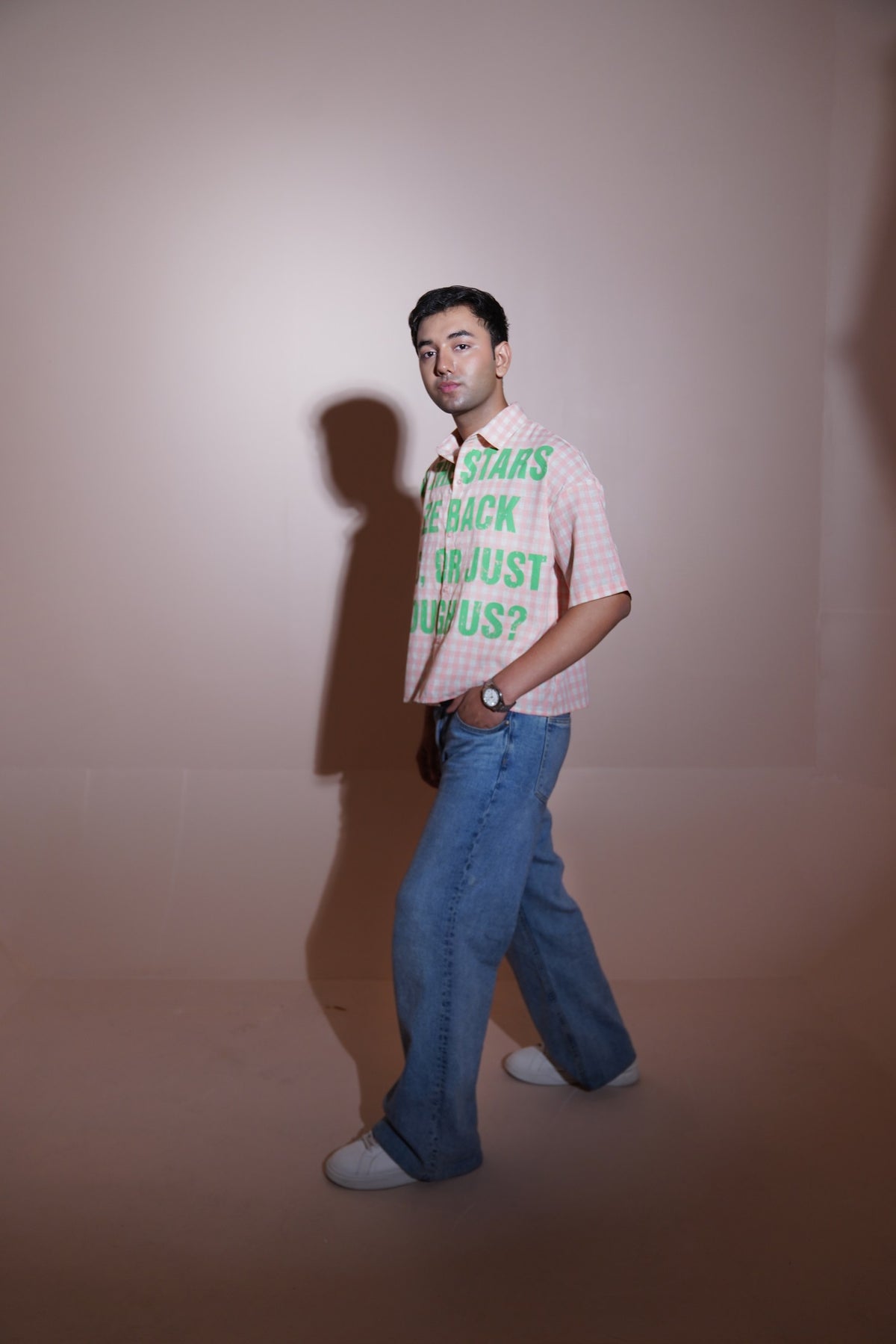 A man wearing a pink and white checkered short-sleeve shirt with green text that reads 'I'M STARS BE BACK OR JUST ABUS?' paired with wide-leg blue jeans and white sneakers, standing against a beige studio backdrop.