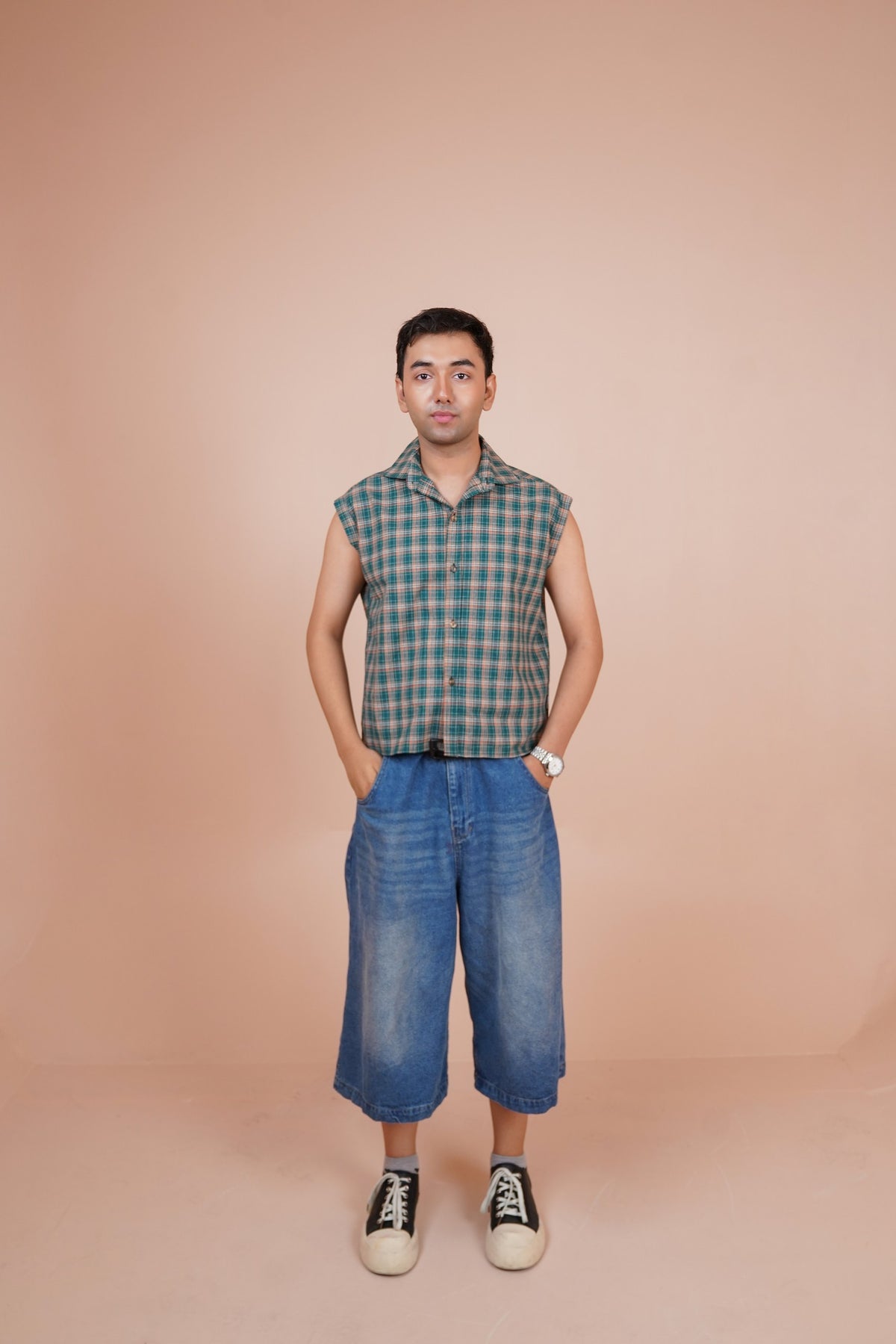 A man stands against a solid peach-colored studio background wearing a sleeveless shirt, blue cropped denim shorts, and black-and-white sneakers.