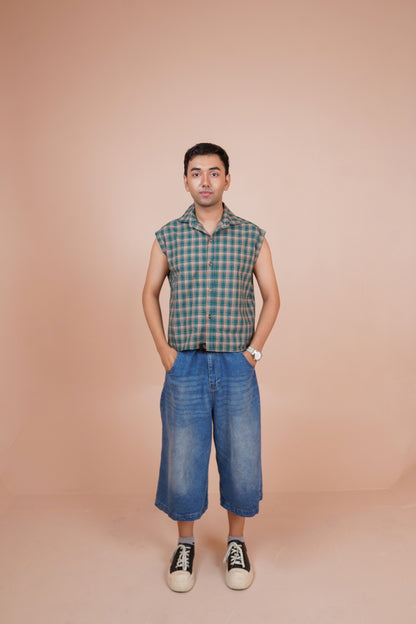 A man stands against a solid peach-colored studio background wearing a sleeveless shirt, blue cropped denim shorts, and black-and-white sneakers.