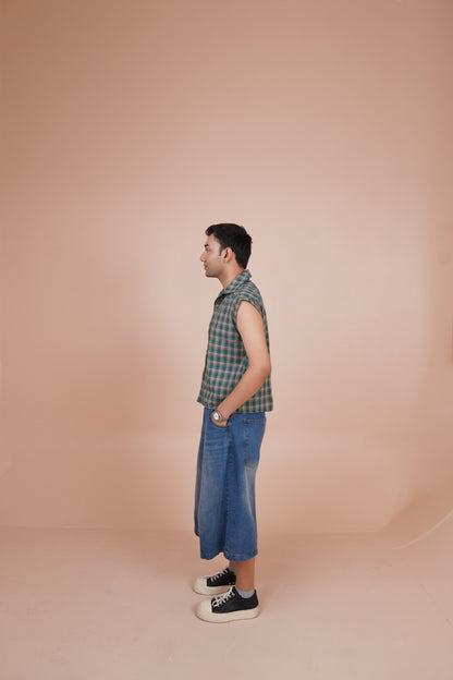 A man stands in profile wearing a sleeveless shirt, blue wide-leg cropped jeans, and black sneakers with white soles, set against a solid dusty pink studio backdrop.