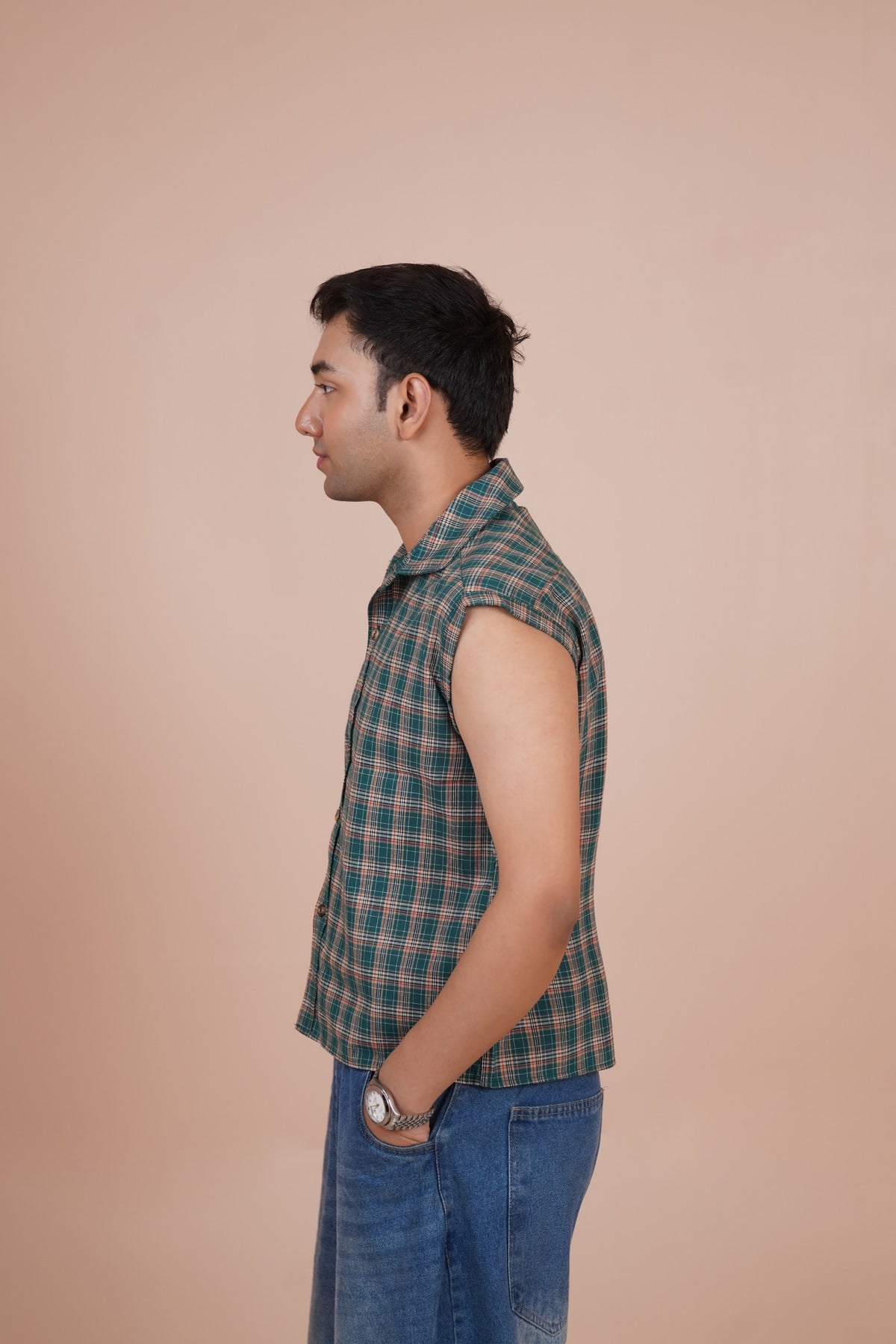 A man shown in profile wearing a sleeveless shirt and blue jeans, standing against a solid beige background.