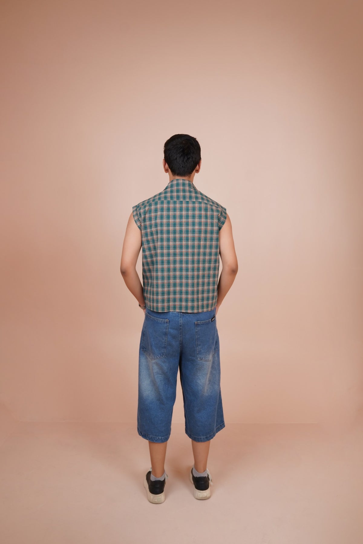 A man viewed from behind wearing a sleeveless shirt and blue denim shorts, standing against a solid peach-colored studio background.