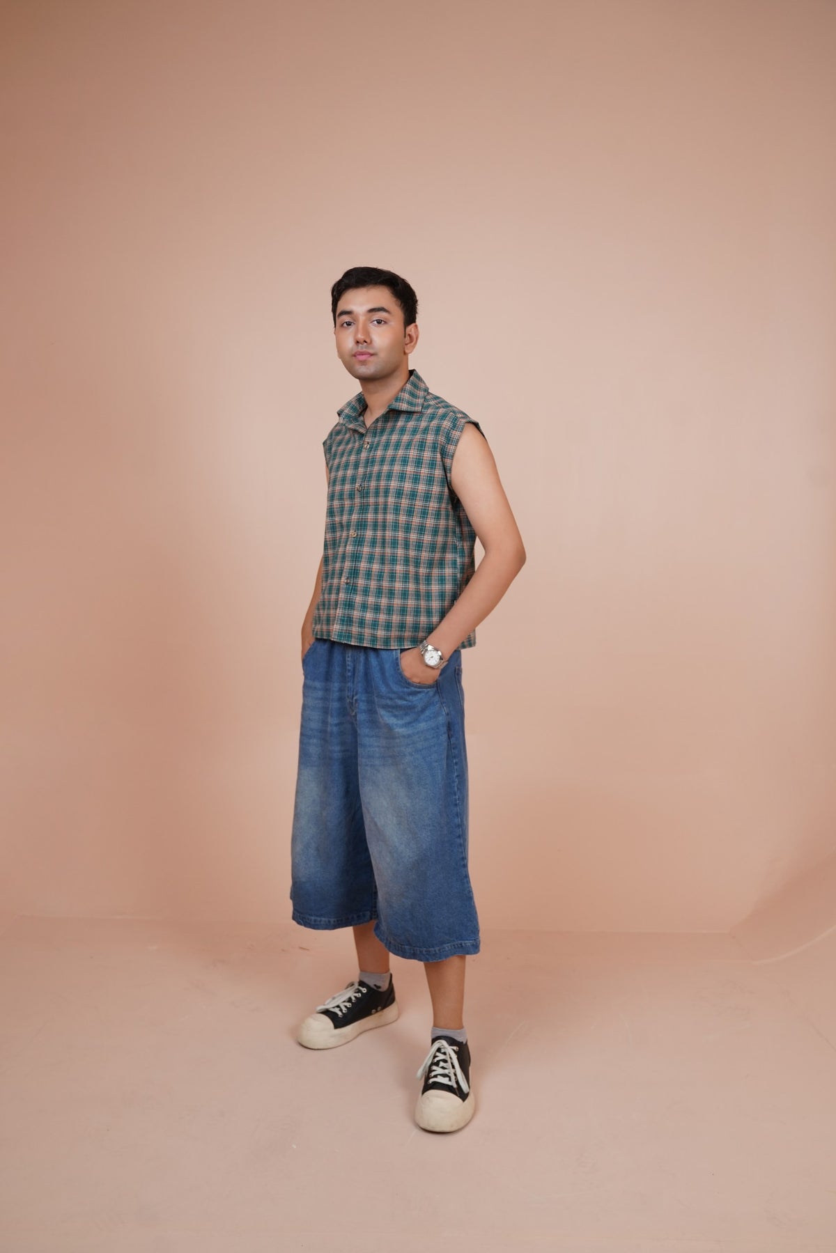 A man modeling a sleeveless shirt paired with wide-leg blue denim shorts and black-and-white sneakers, standing against a solid peach-pink studio backdrop.