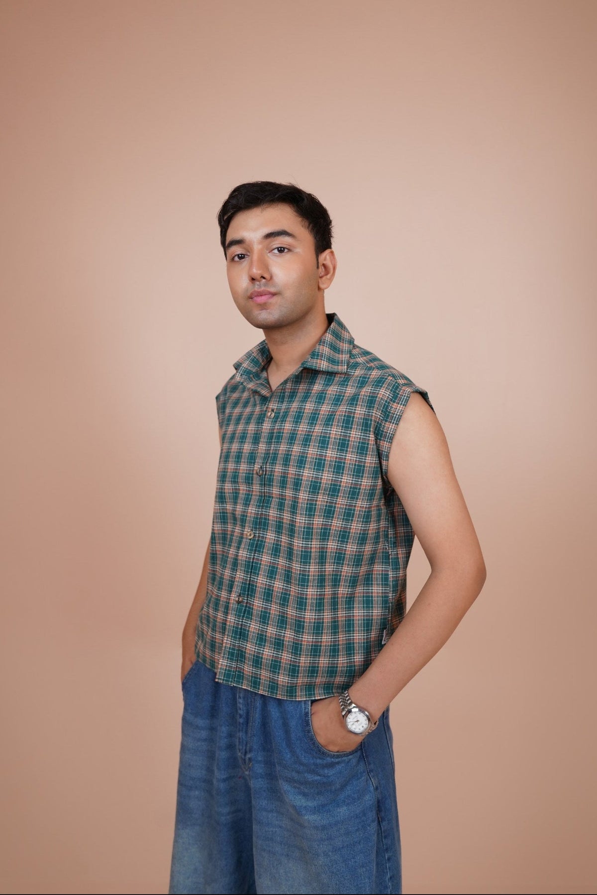 A man wearing a sleeveless shirt and blue jeans, standing against a solid beige background.
