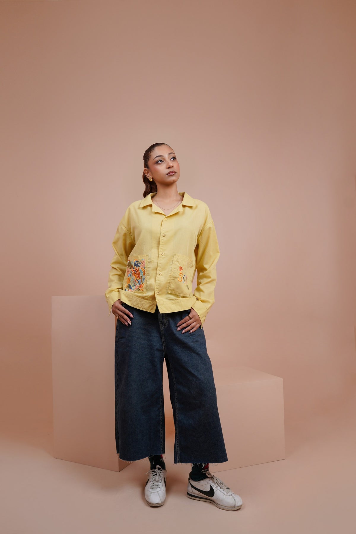 A model stands in a studio setting wearing a Yellow long sleeve button up shirt with colorful floral embroidery on the front, paired with dark wide-leg pants and white sneakers, against a solid peach-pink background.