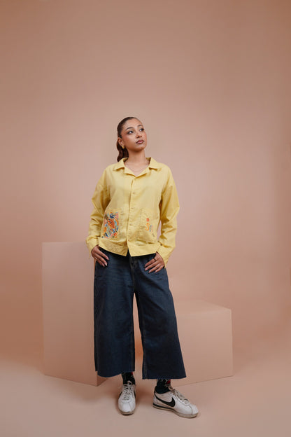 A model stands in a studio setting wearing a Yellow long sleeve button up shirt with colorful floral embroidery on the front, paired with dark wide-leg pants and white sneakers, against a solid peach-pink background.