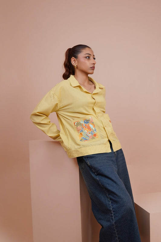 A woman wearing a pale Yellow long-sleeve button-up shirt with a colorful embroidered patch on the pocket, paired with dark blue jeans, posing against a solid dusty pink background.