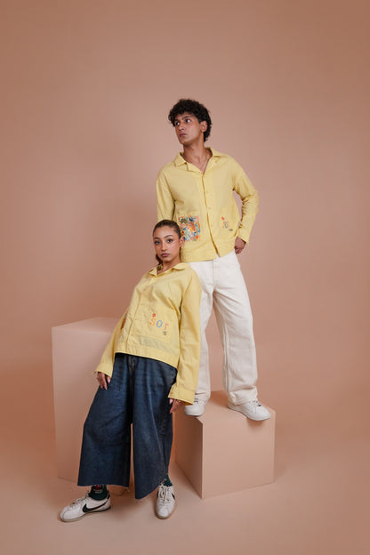 Two models posing in matching pale Yellow long sleeve button up shirt, one standing on a pink block and one sitting on a pink block against a solid pink background.
