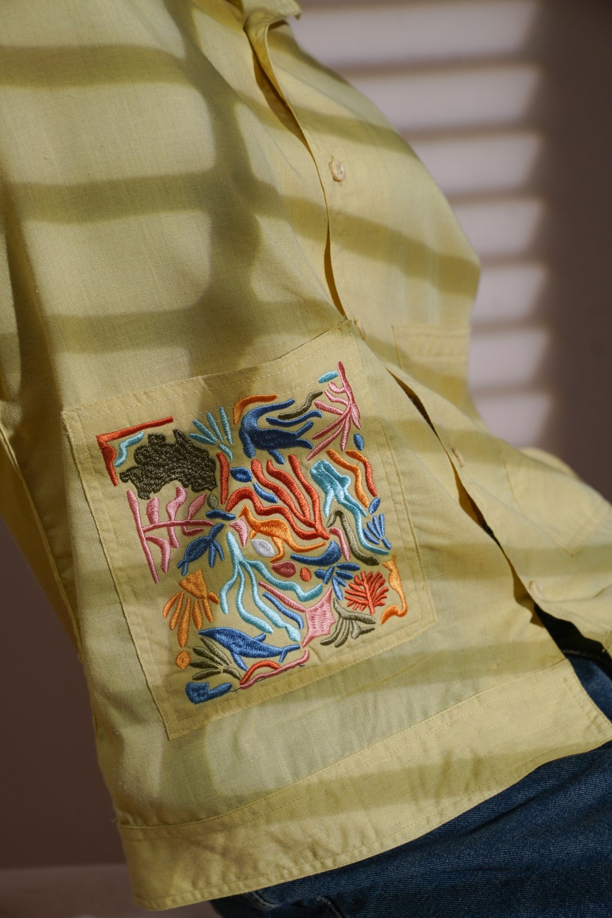 A close-up of a pale Yellow long-sleeve button-up shirt worn by a person, featuring a square embroidered patch on the chest pocket with colorful floral and leaf motifs in blue, orange, pink, and green.