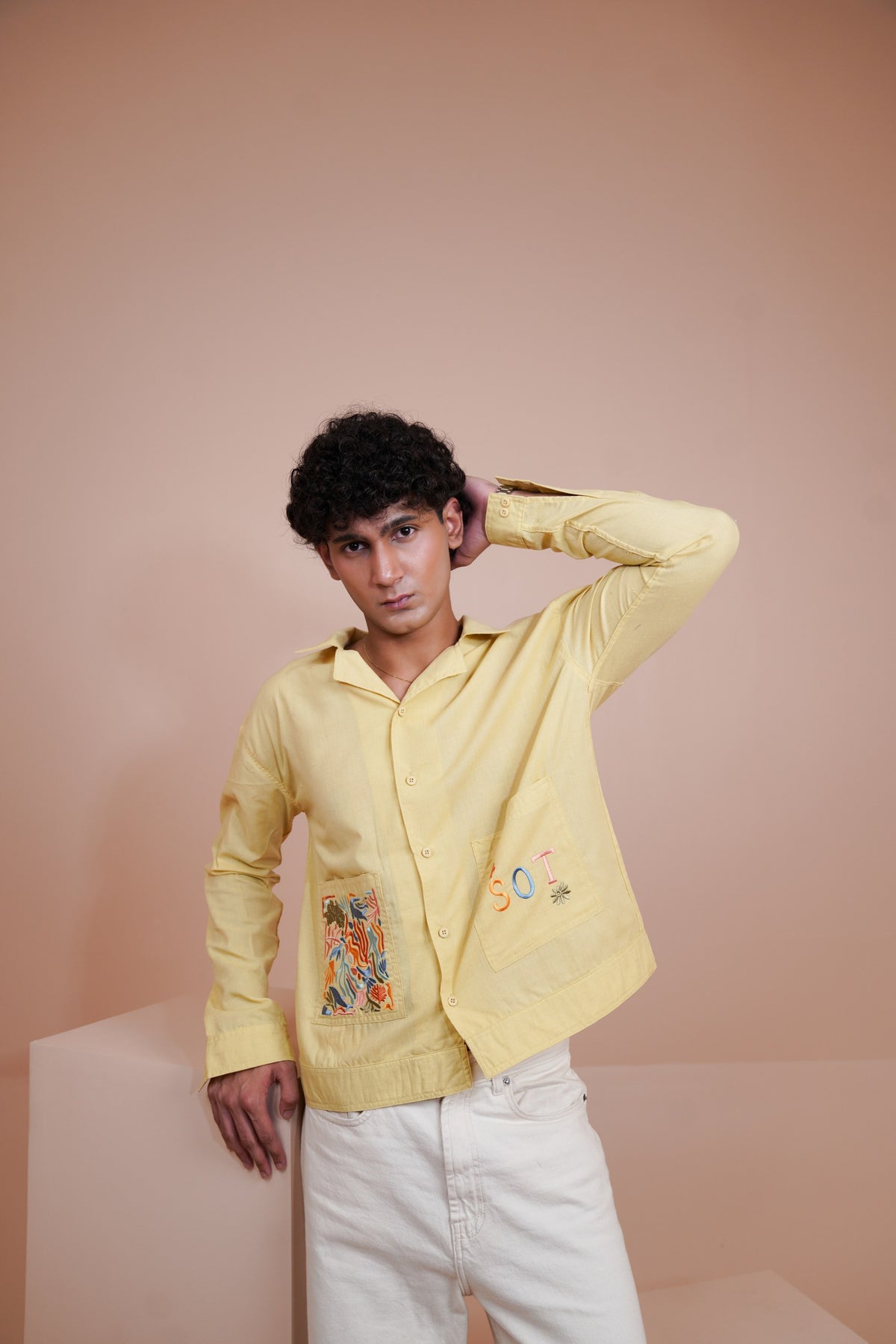 A man wearing a pale Yellow long sleeve button up shirt with colorful embroidered details on the front pockets, paired with white pants, posing against a solid peach-pink studio background.
