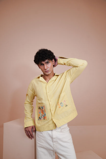 A man wearing a pale Yellow long sleeve button up shirt with colorful embroidered details on the front pockets, paired with white pants, posing against a solid peach-pink studio background.