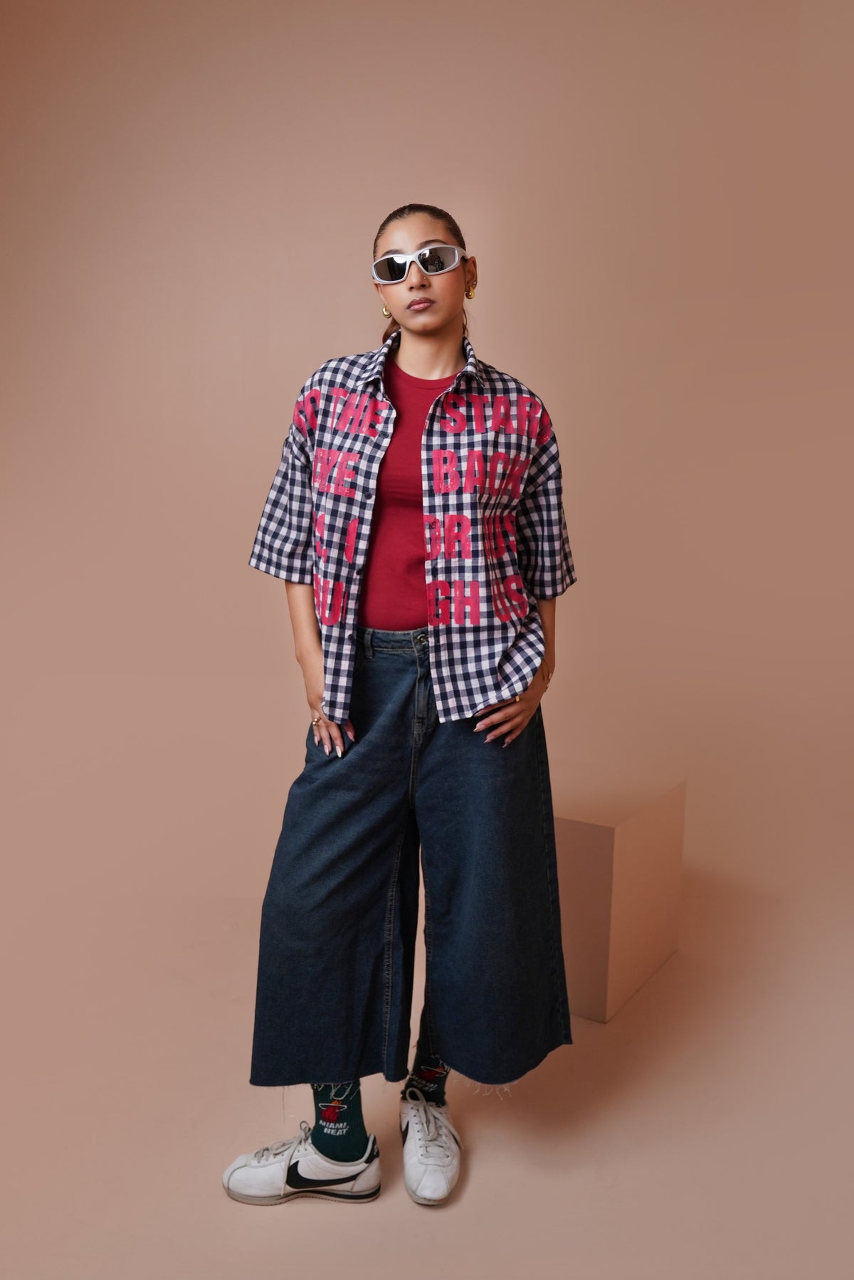 A model stands against a solid warm beige studio background wearing a short-sleeve gingham unisex streetwear shirt with pink graphic text, a red t-shirt, wide-leg dark denim jeans, patterned socks, and white sneakers.