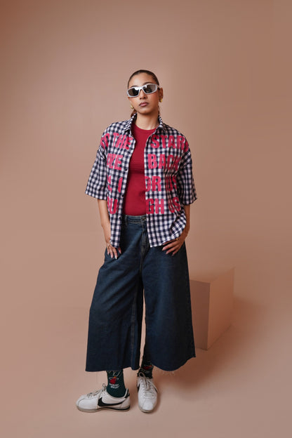 A model stands against a solid warm beige studio background wearing a short-sleeve gingham unisex streetwear shirt with pink graphic text, a red t-shirt, wide-leg dark denim jeans, patterned socks, and white sneakers.