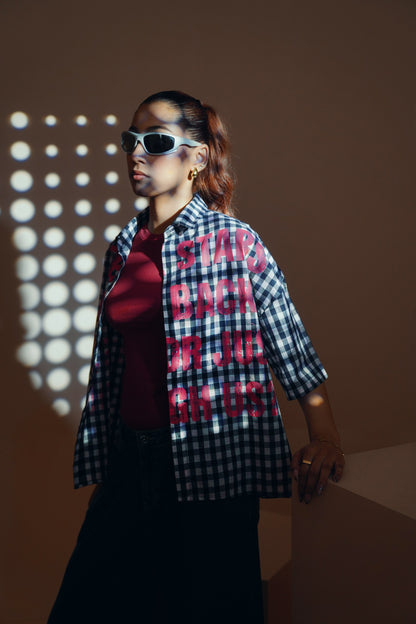 A woman wearing a black and white gingham unisex streetwear shirt with bold red text reading 'STAY BACK OR DIE ON US' over a dark red top and black pants, styled with white sunglasses and gold hoop earrings in a warm-toned studio setting with projected light patterns.