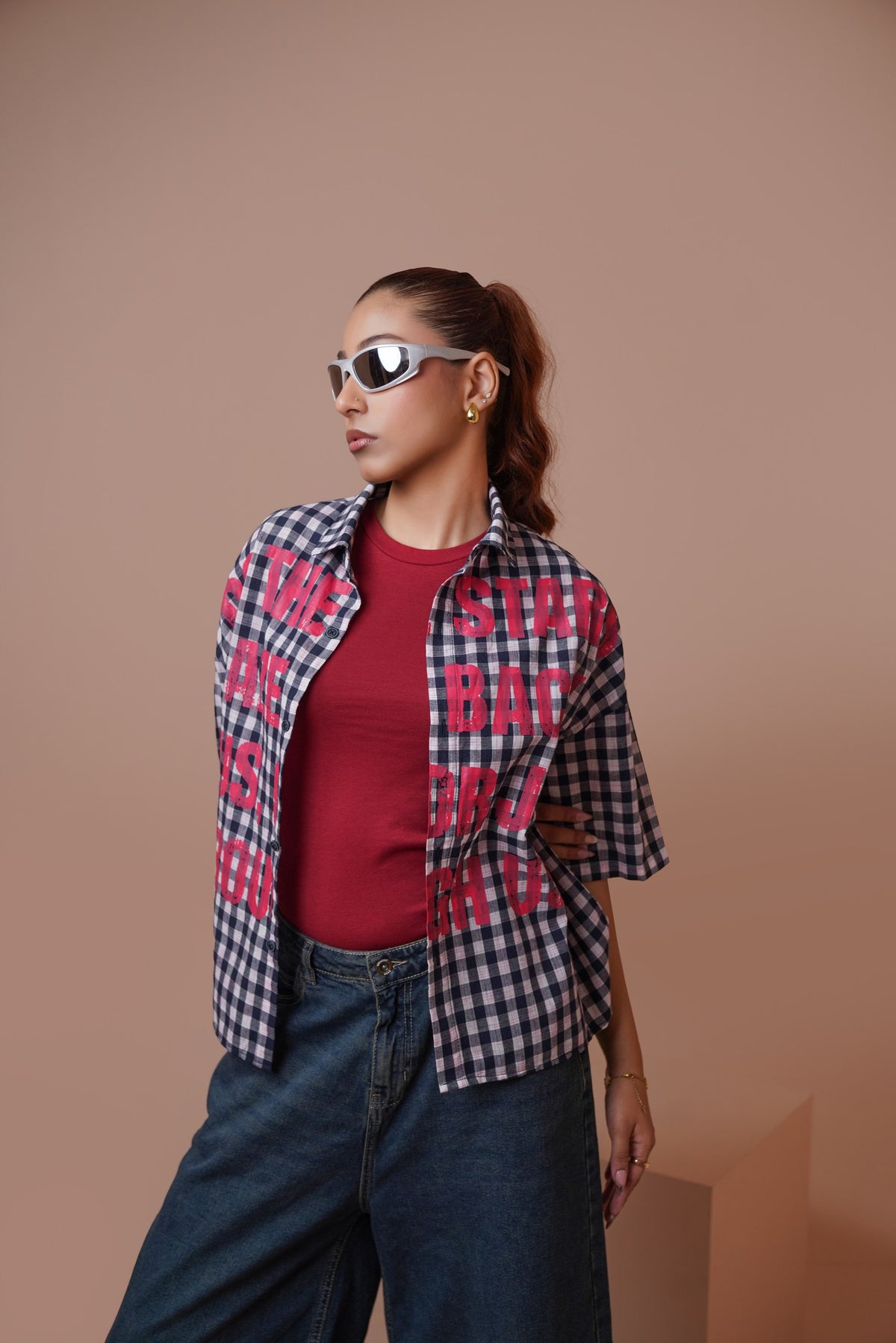 A woman modeling a short-sleeve plaid unisex streetwear shirt with bold pink text graphics over a red t-shirt and blue jeans, set against a solid warm beige studio background.