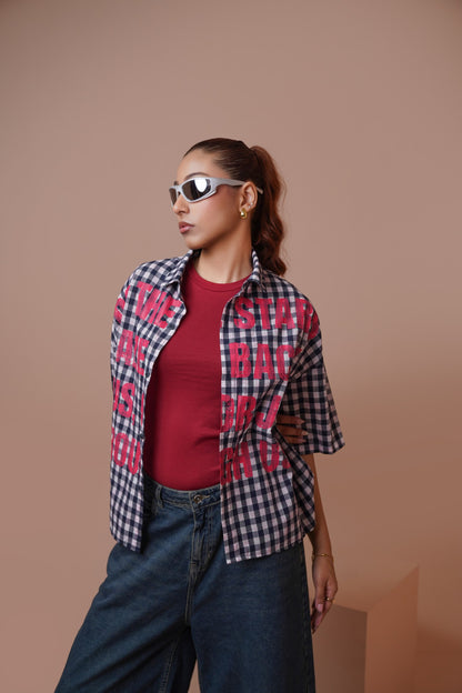 A woman modeling a short-sleeve plaid unisex streetwear shirt with bold pink text graphics over a red t-shirt and blue jeans, set against a solid warm beige studio background.