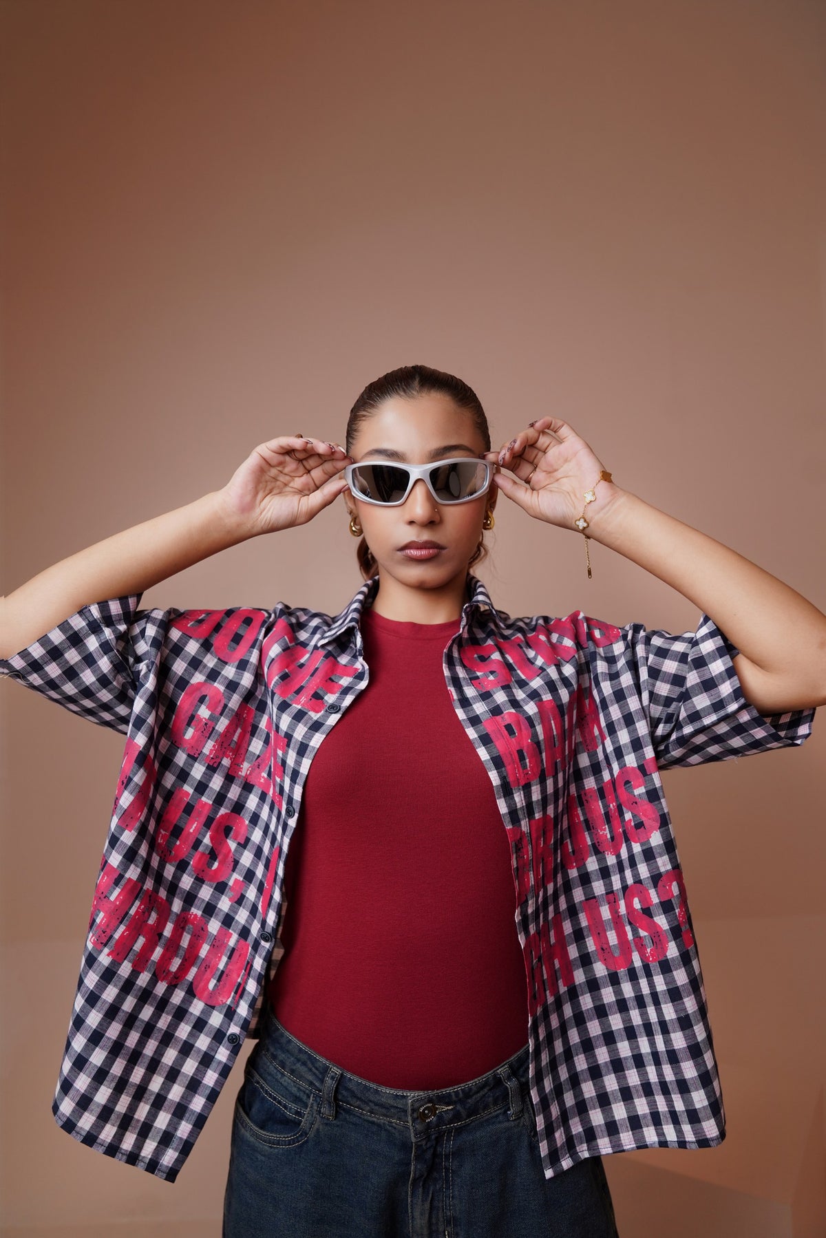 A model wearing a black and white gingham unisex streetwear shirt with bold pink text graphics, layered over a red ribbed top and blue jeans, posing against a solid warm brown background.