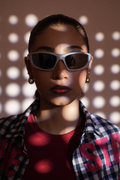 A close-up portrait of a model wearing white-framed cat-eye sunglasses with dark lenses, a red t-shirt, and a black-and-white checkered flannel shirt. The background is a warm brown wall with circular light patterns casting shadows.