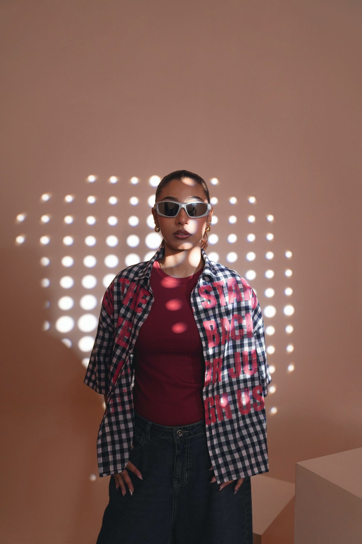 A model wearing a red t-shirt, a black and white gingham overshirt with red graphic text, and dark jeans, standing against a warm beige wall with circular light projections.