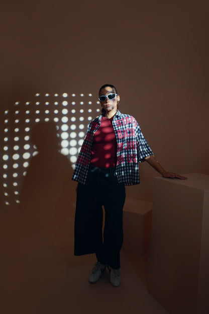 A model wearing a red and black plaid short-sleeve unisex streetwear shirt over a red t-shirt, paired with black wide-leg pants and white sneakers, standing in a minimalist studio with geometric blocks and light spot shadows on a warm brown background.