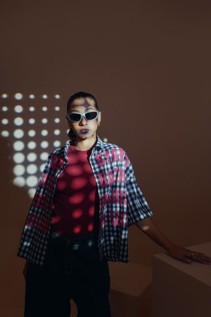A model wearing a red t-shirt, a black and white plaid unisex streetwear shirt, and black pants, standing in a studio with dramatic light projections on a brown background.