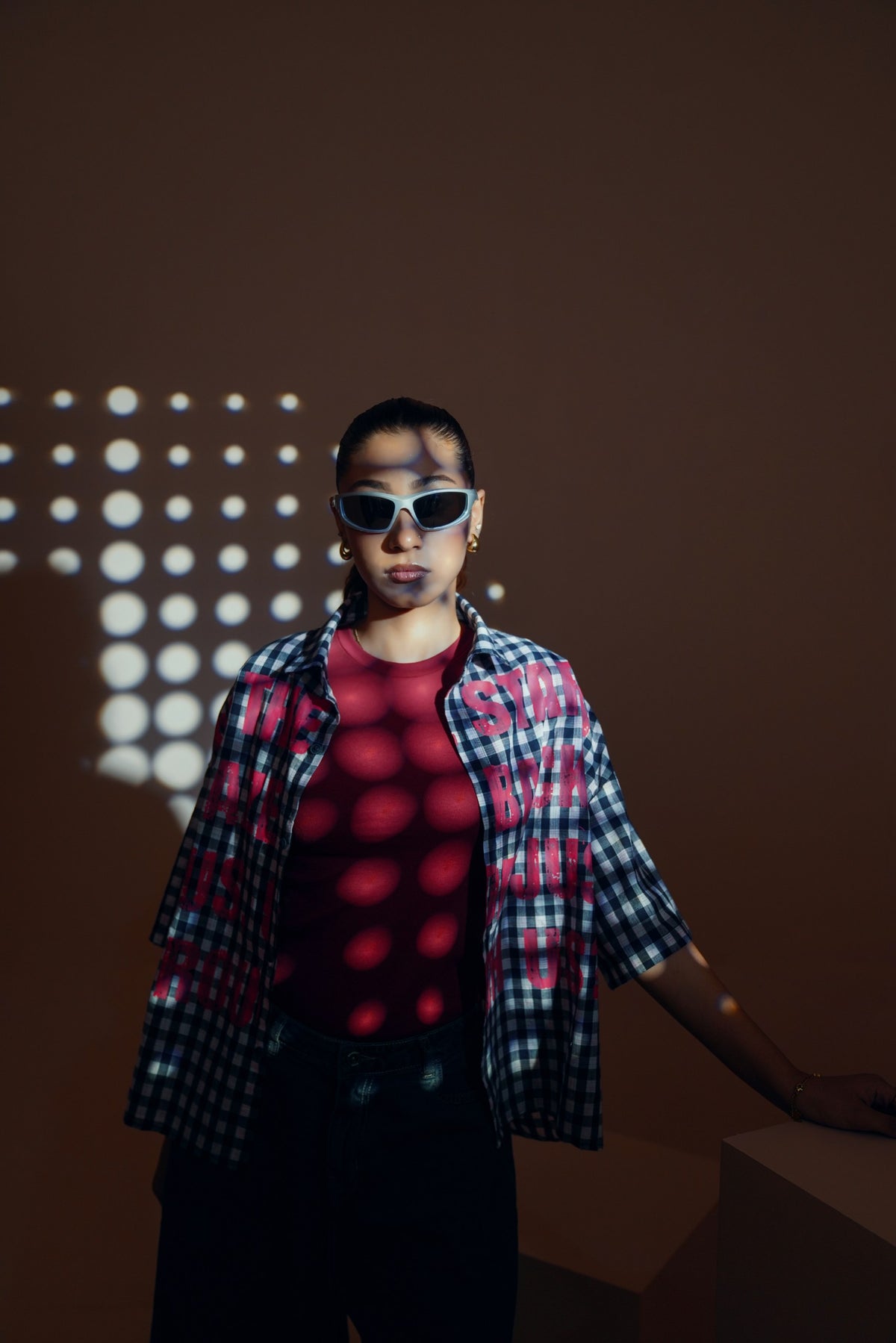 A person wearing white-framed sunglasses, a red t-shirt, a red-and-black plaid overshirt, and black pants, standing against a dark brown wall with circular light projections.