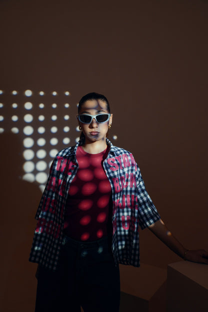 A person wearing white-framed sunglasses, a red t-shirt, a red-and-black plaid overshirt, and black pants, standing against a dark brown wall with circular light projections.
